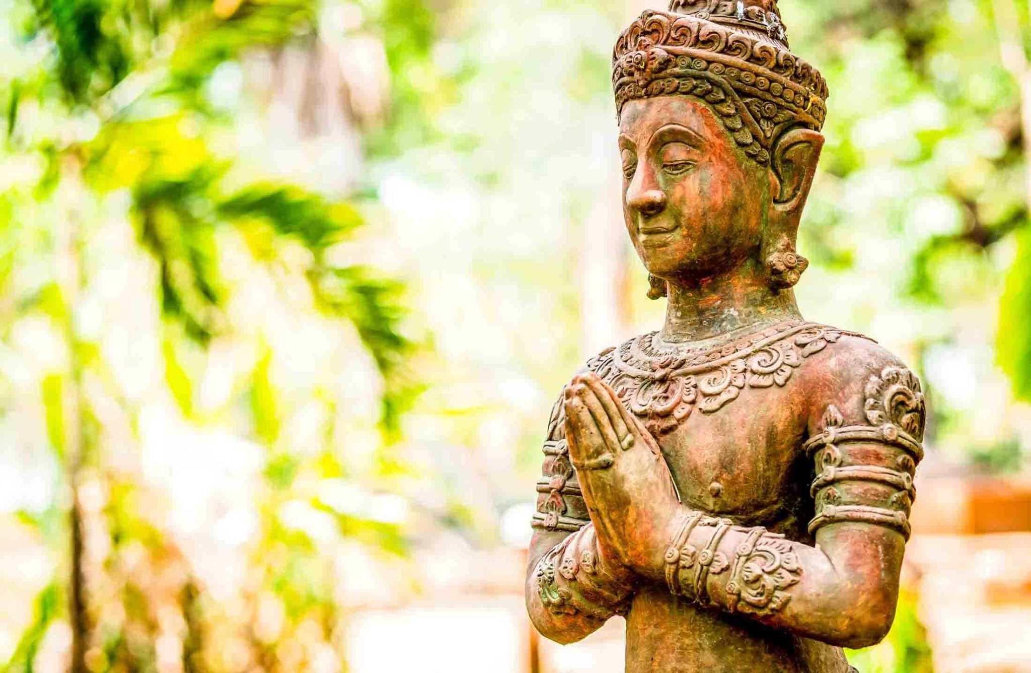 Buddha statue with hands in prayer position amidst lush greenery.